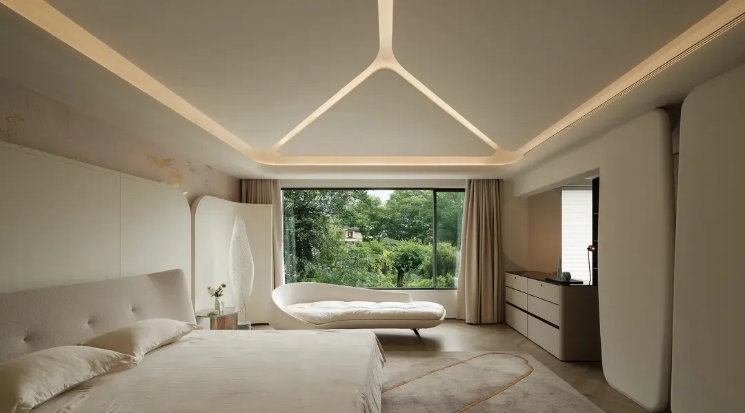 A modern bedroom with neutral colors, including a bed with a curved headboard, a white lounge chair, and a large window with