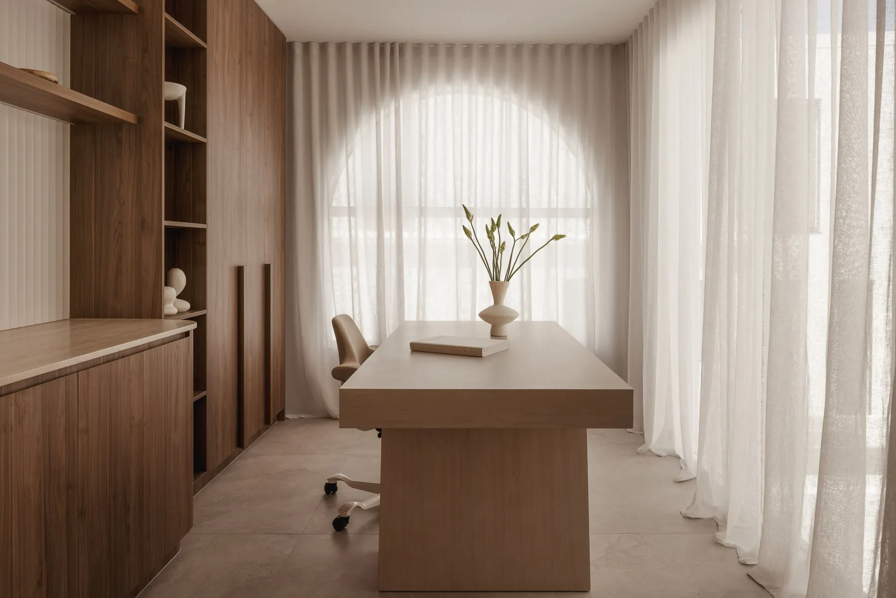 A modern minimalist office space featuring a wooden desk, white fabric curtains, and a vase with green plants.