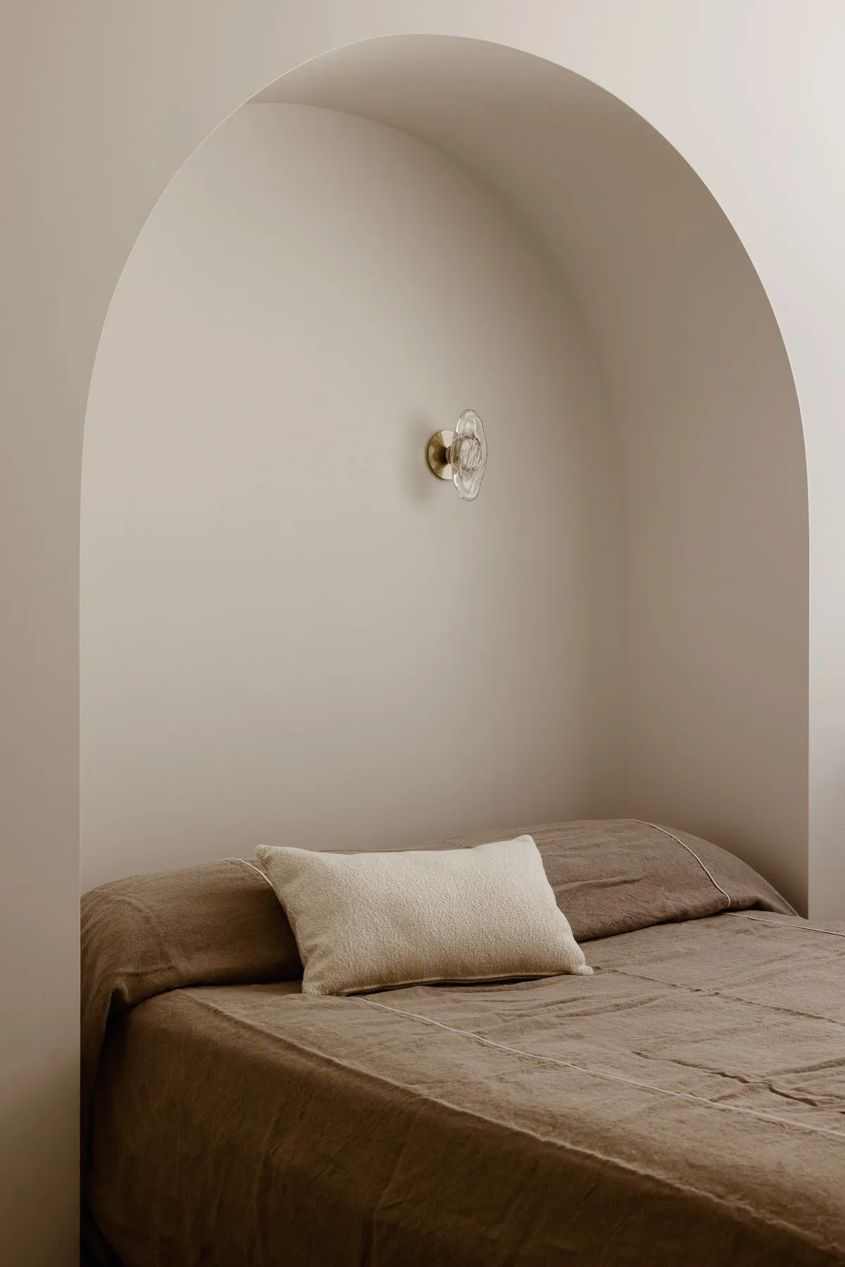 A minimalist modern bedroom with a bed covered in neutral textiles, framed by an archway and a sconce fixture.