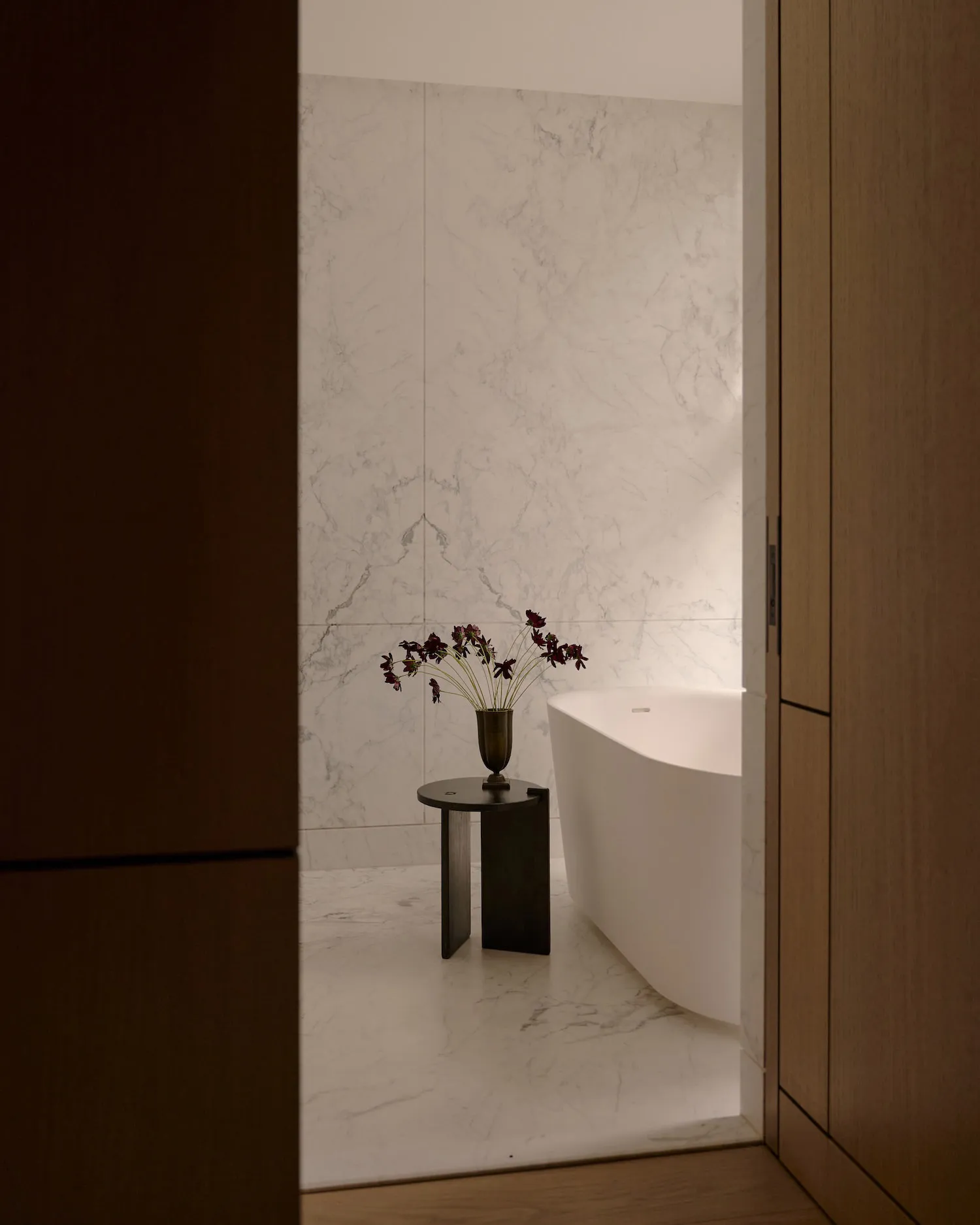 Modern minimalist bathroom featuring a white marble bathtub and a small table with a vase of flowers.