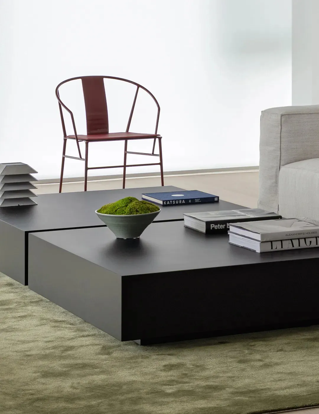 A modern living room featuring a black metal platform coffee table, a grey sofa, a red chair, and a green vase.