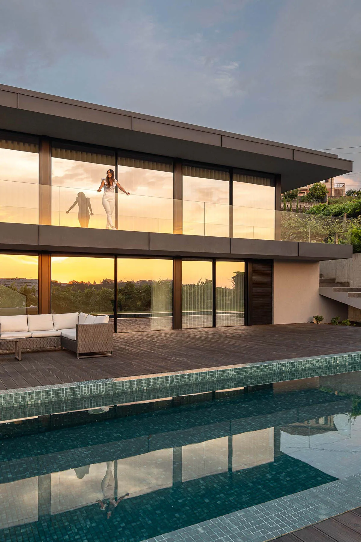 Modern residential architecture featuring glass walls and wooden deck with a swimming pool and sunset view.