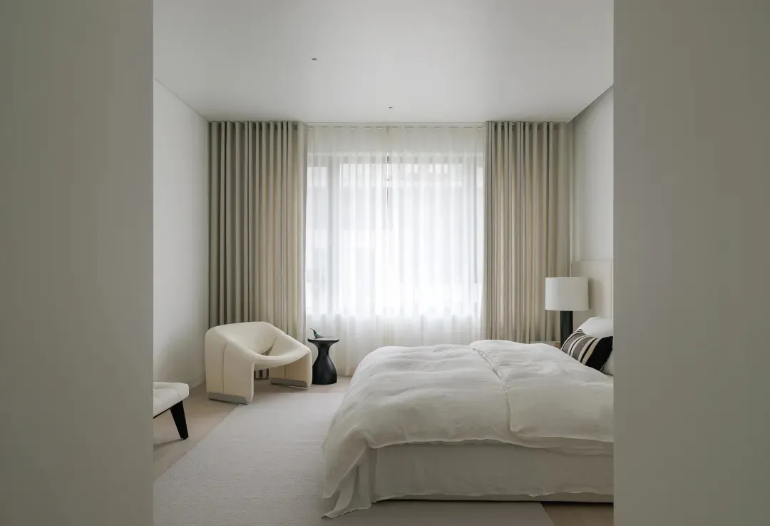 A minimalist bedroom featuring neutral-colored furniture and curtains, large windows with white sheer curtains, and a white b