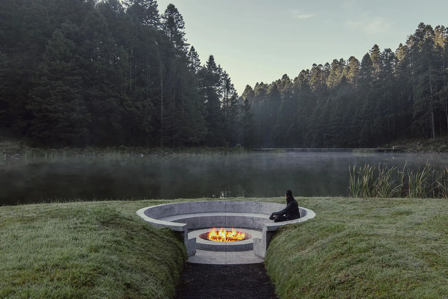 Concrete firepit surrounded by grass with a person sitting next to it in a serene forested landscape.