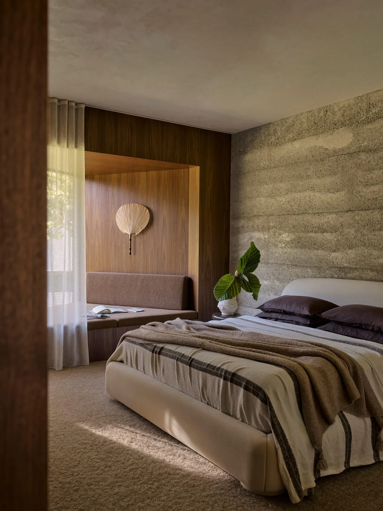 A minimalist bedroom featuring concrete and wood materials, a white bed with a striped blanket, and a wooden bench against th