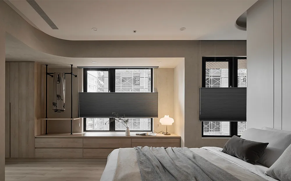 A modern minimalist bedroom with light wood flooring, a white bed with grey throw blankets, and a wooden cabinet with open sh