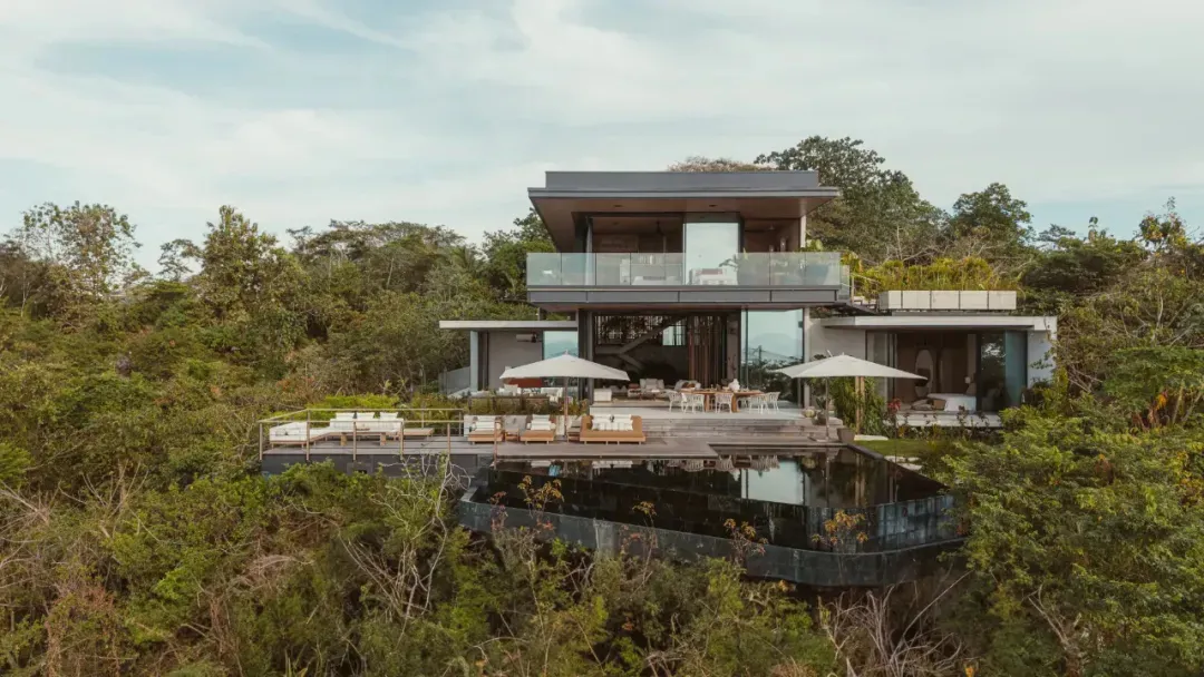 Modern vacation home with glass and concrete exterior, surrounded by lush greenery.