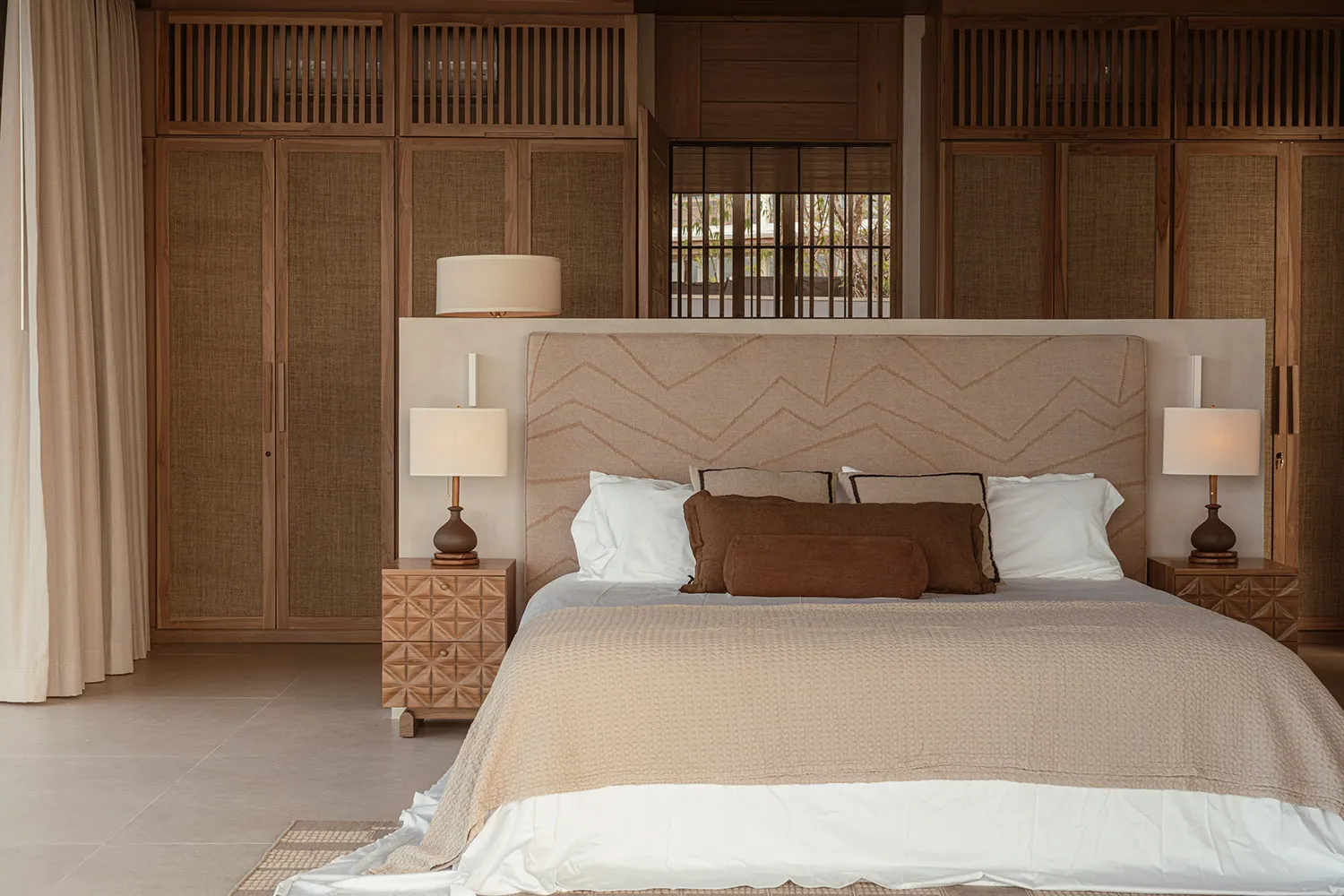 A minimalist bedroom featuring a taupe upholstered bed with matching comforter, surrounded by wooden furniture and a large wi