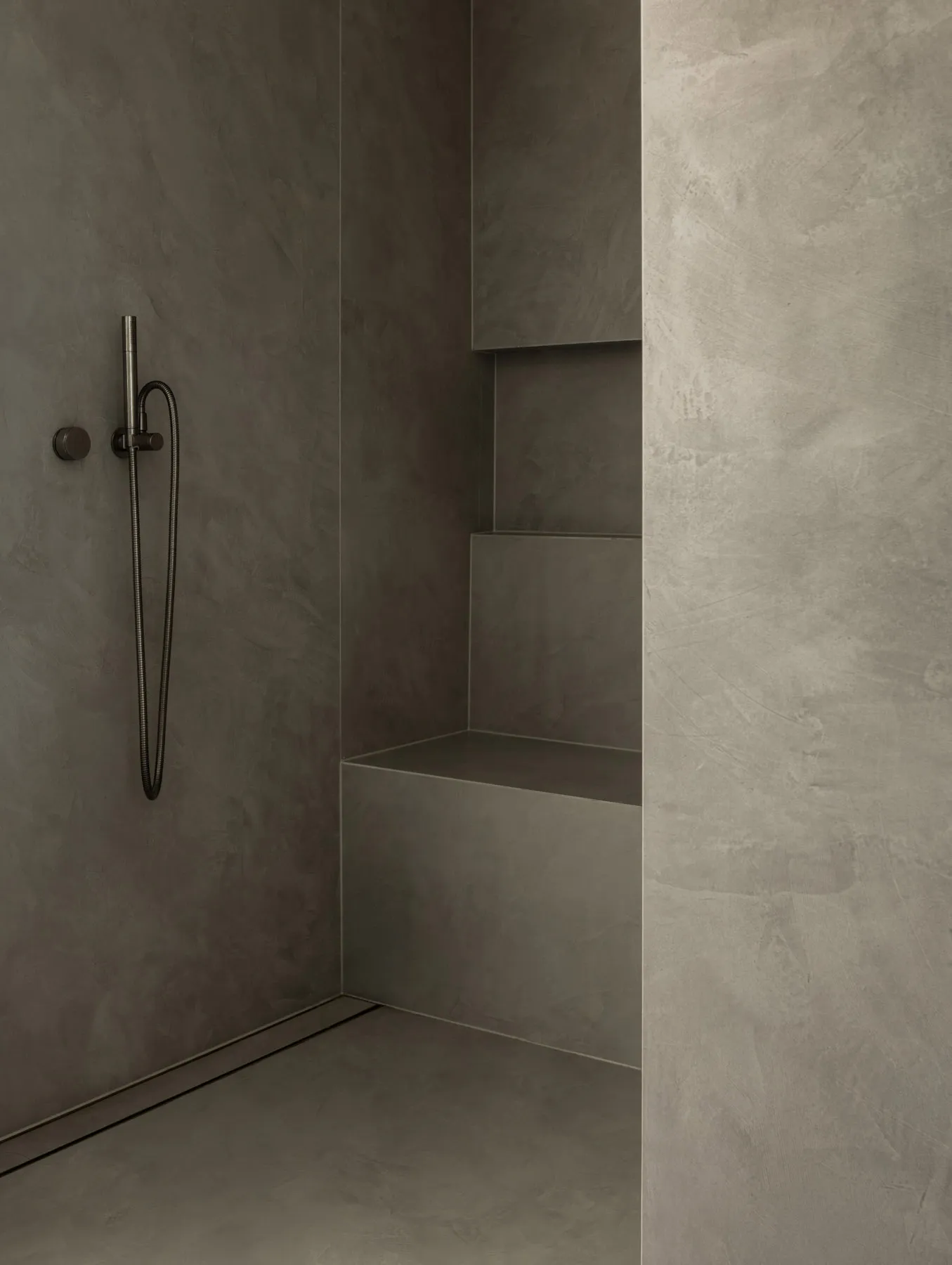 A minimalist bathroom with concrete walls and floors, featuring a modern shower area with a hand shower head and built-in she
