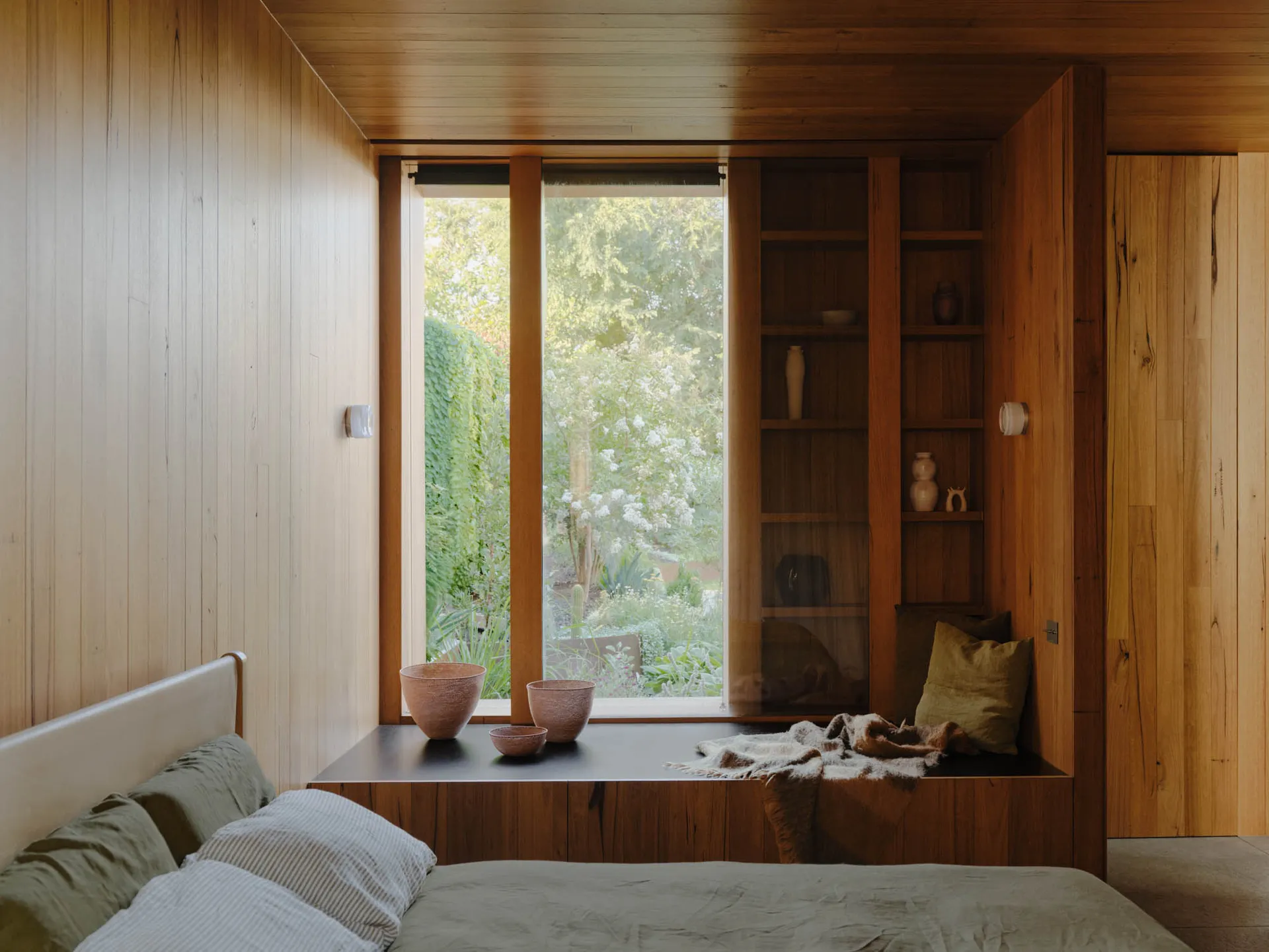 Bedroom with large windows showcasing lush greenery outside, wooden panel walls, and a bench seat with bowls and a blanket.