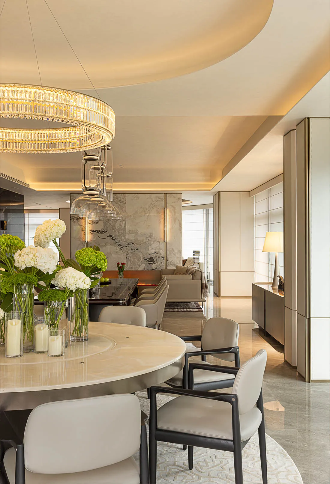 A modern residential dining room featuring a round marble dining table, white leather chairs, and a crystal chandelier.