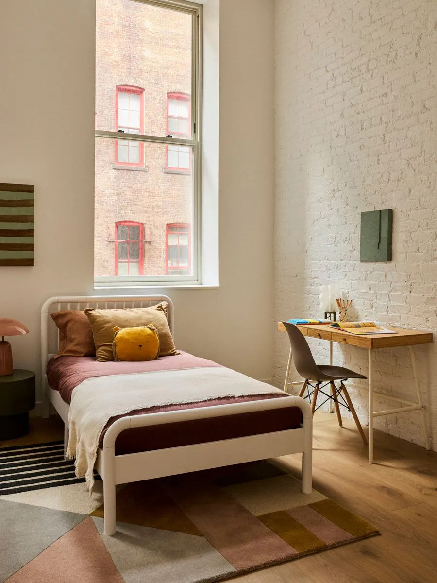 A minimalist bedroom featuring a white bed, colorful pillows, and a wooden desk against a white brick wall.