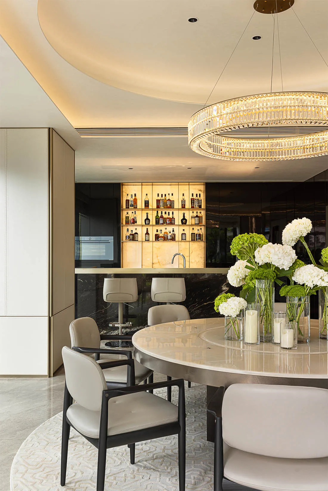 A modern dining room with a round marble table surrounded by beige chairs, a bar with illuminated bottles, and a crystal chan