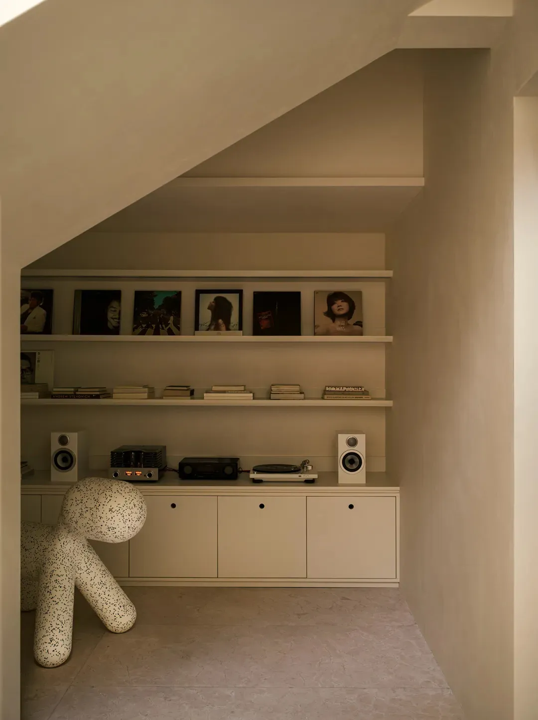 A modern music room with a concrete floor, shelves displaying framed album covers and books, and a white audio speaker.