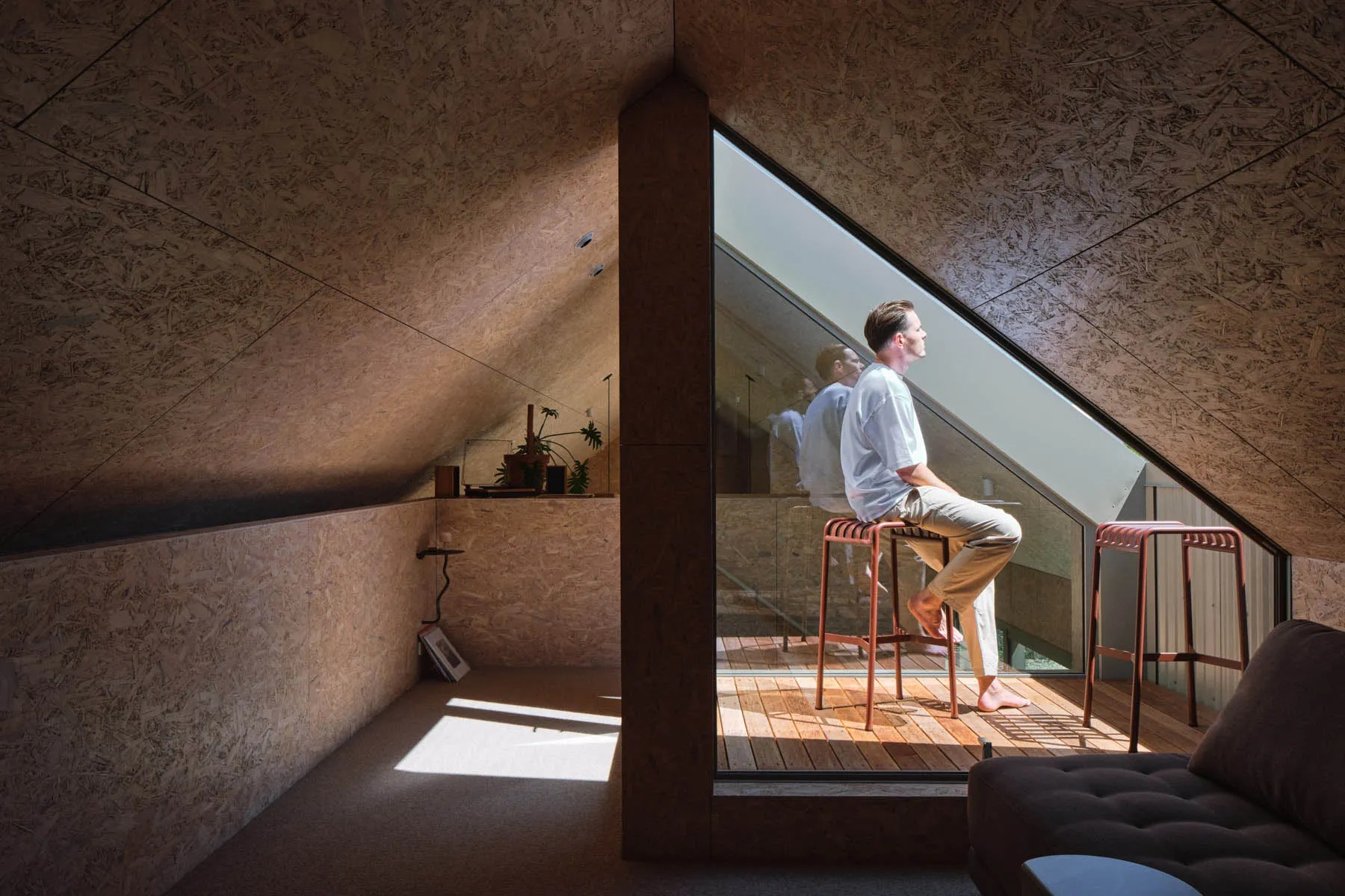 A minimalist living space with wooden floors and a person sitting on a stool looking out through a large window.