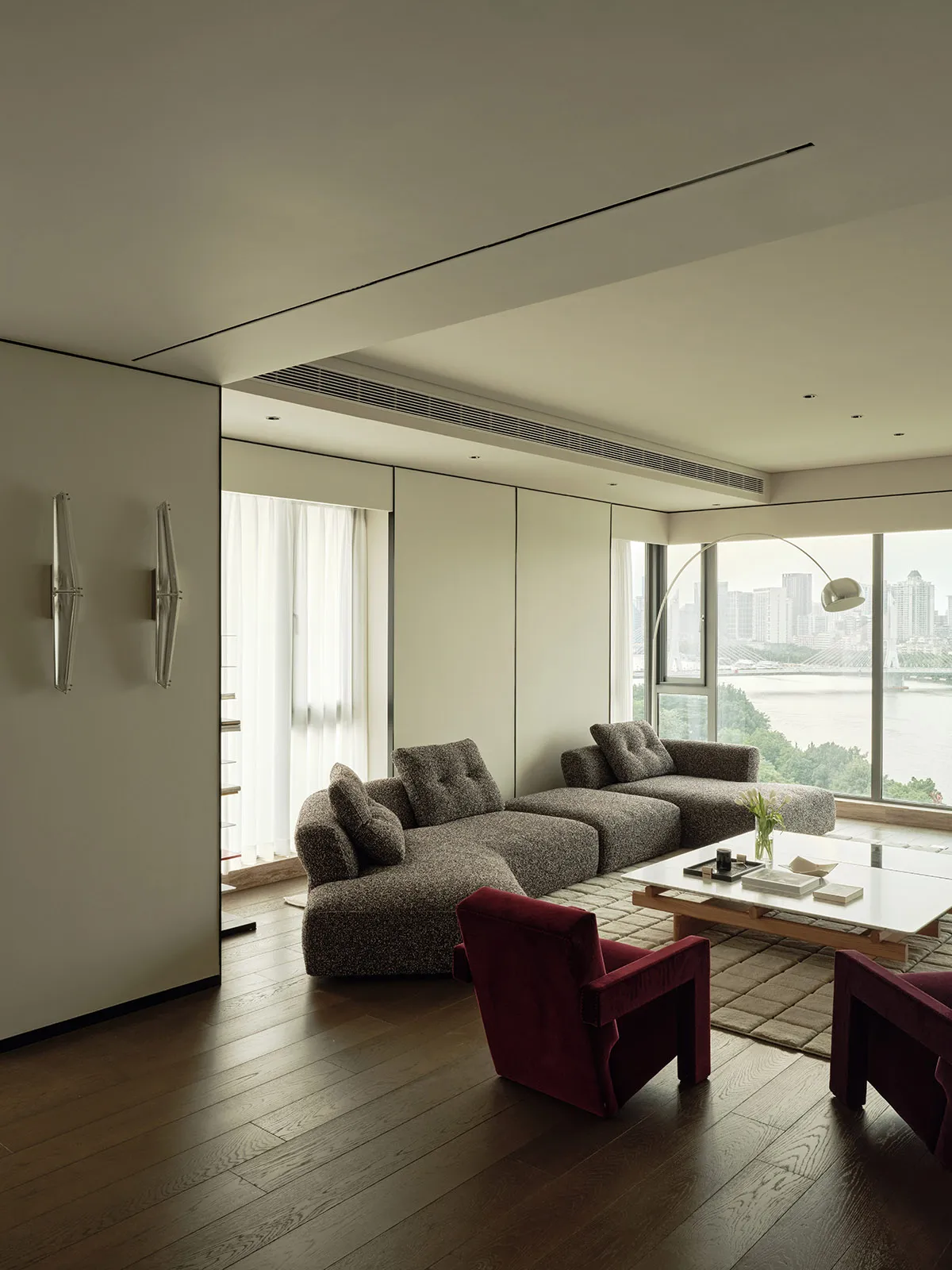A modern living room featuring a fabric sectional sofa, glass windows, and wooden flooring with a ceiling light fixture.