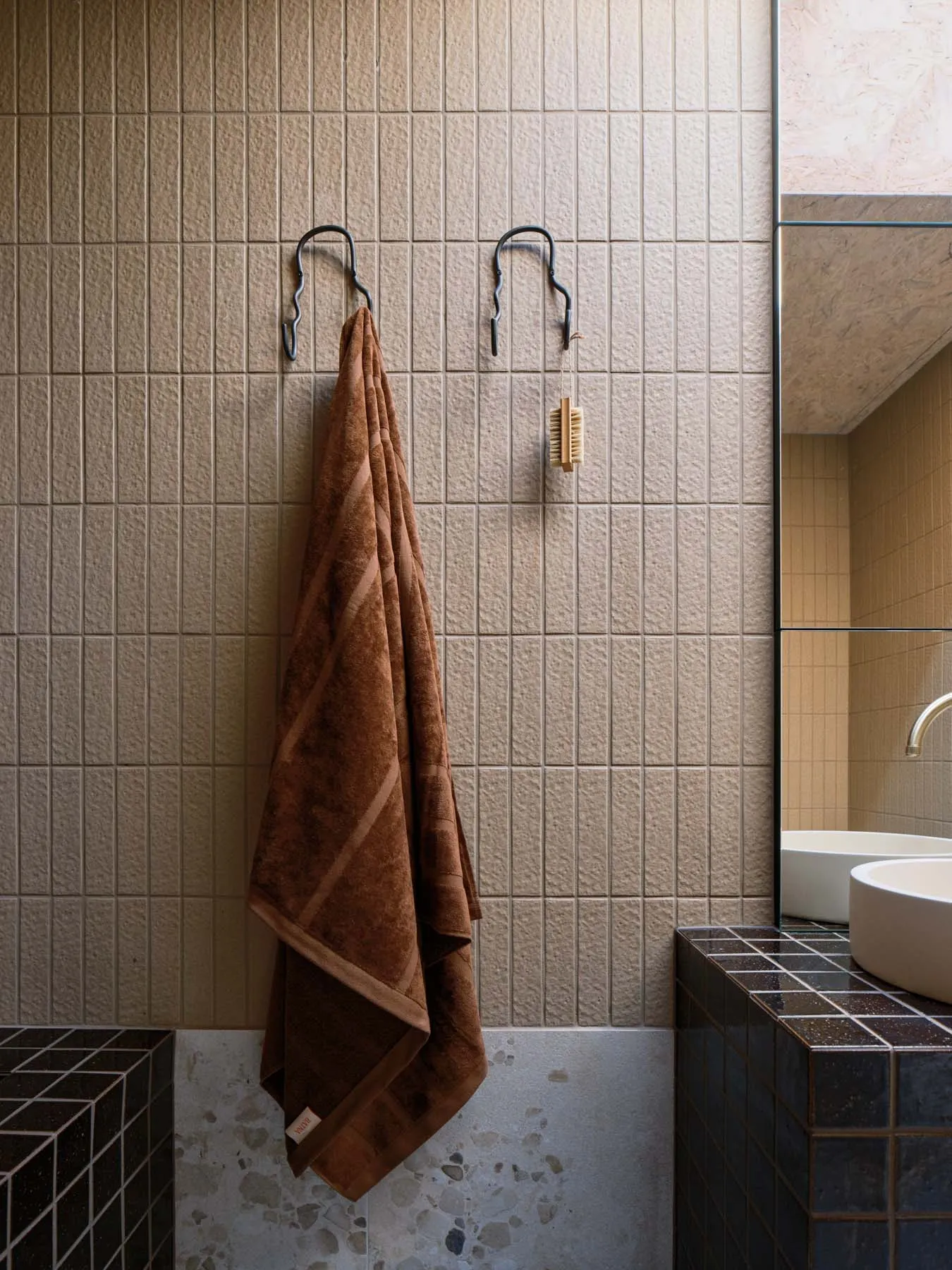 Modern bathroom featuring a textured tile wall with two metal hooks holding a brown towel and a scrubbing brush.