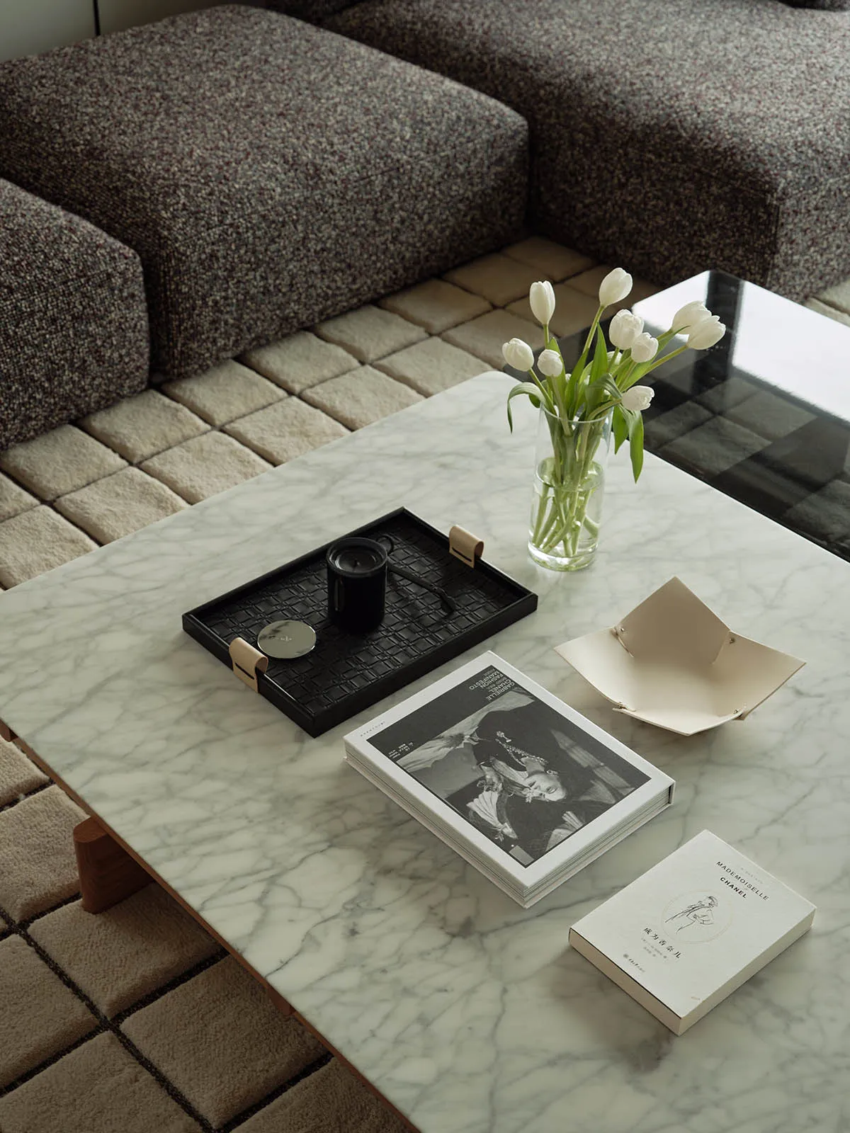 A modern living room featuring a marble coffee table with a black tray, a vase of white tulips, and a book.
