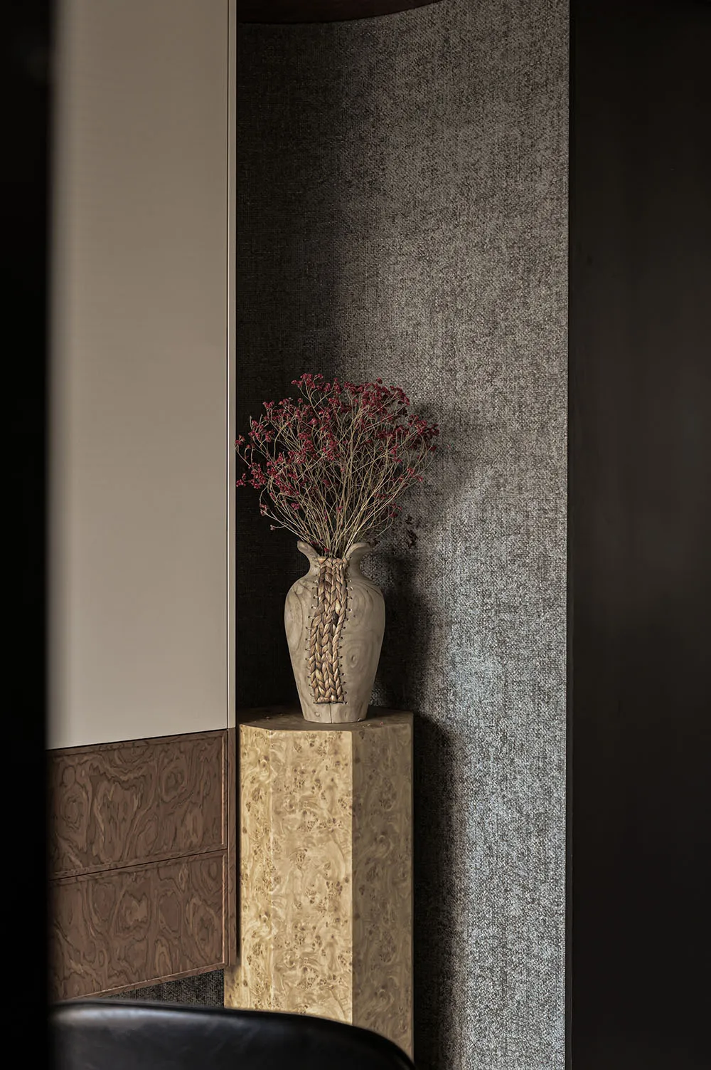 A wooden vase with dried flowers placed on a wooden standing base against a textured grey wall.