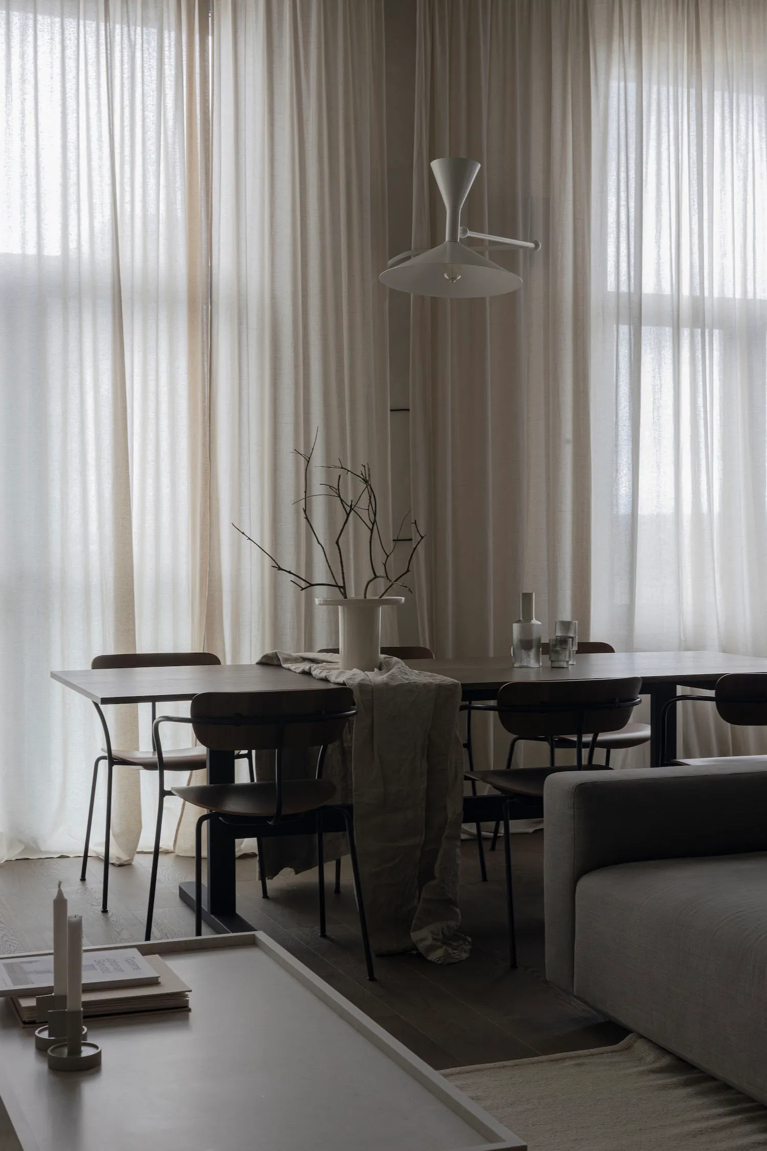 A minimalist residential setting featuring a wooden table, a white lamp, and sheer curtains, with a cozy ambiance.