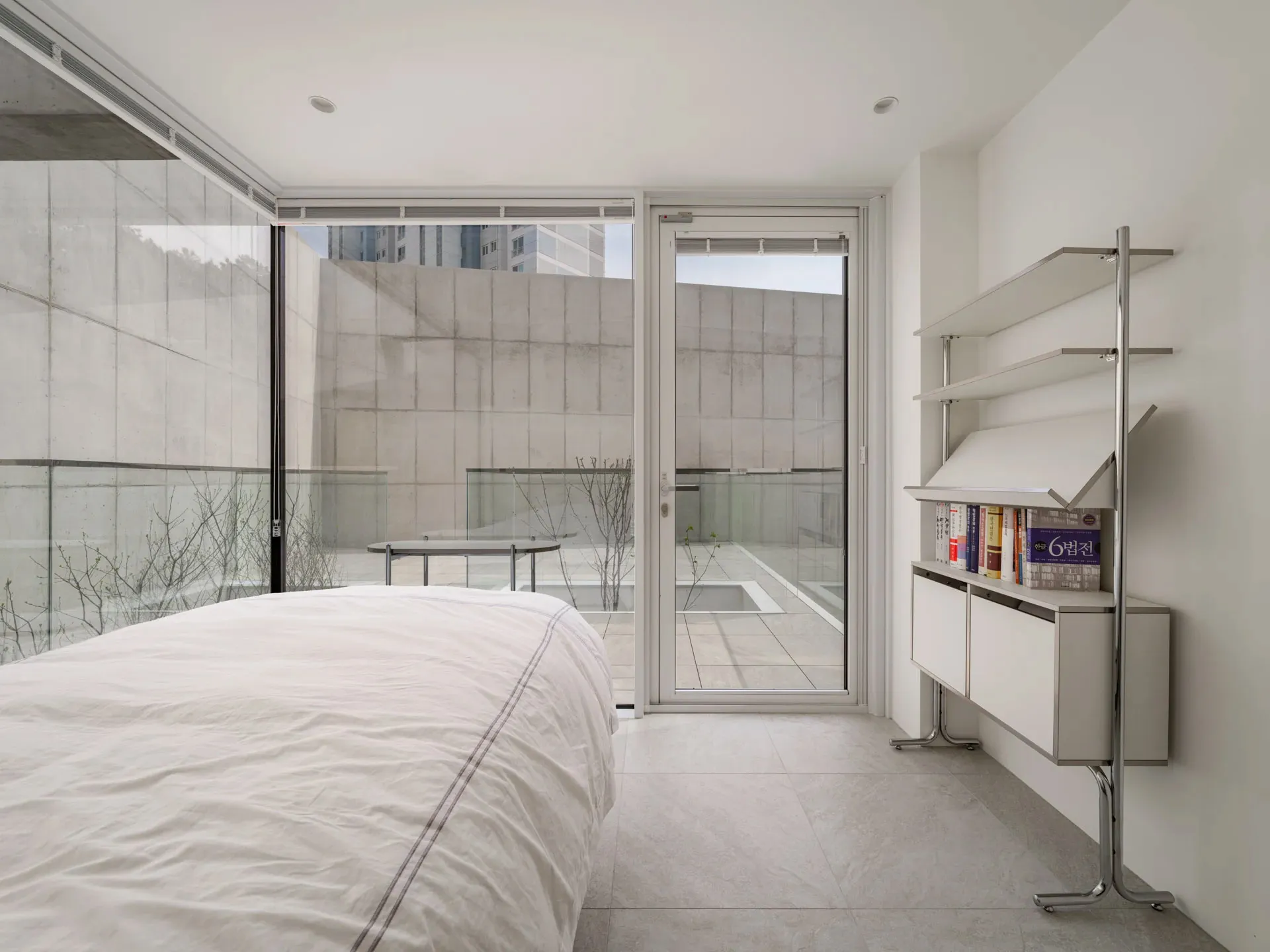 A modern minimalist bedroom with concrete walls, a large bed, and a bookshelf.