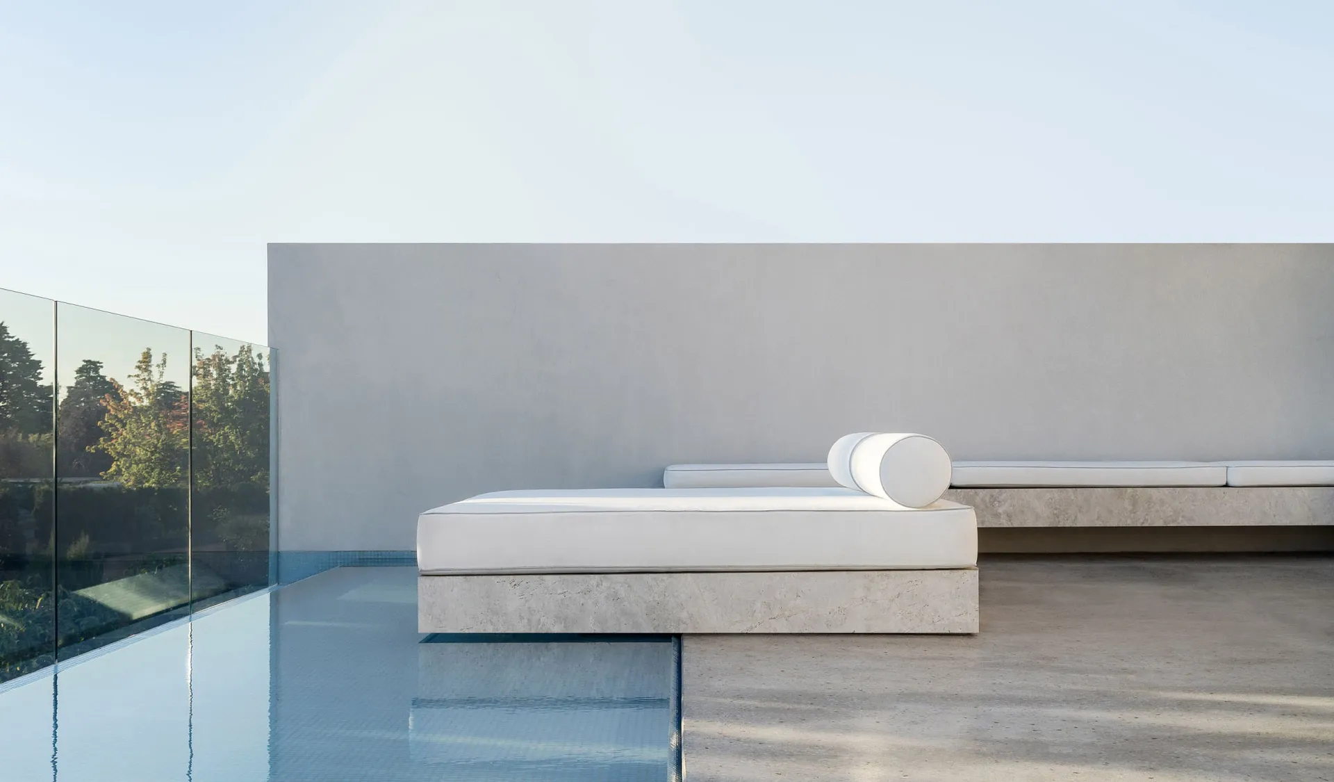Modern, minimalist concrete seating area by a pool with white cushions and a cylindrical pillow.