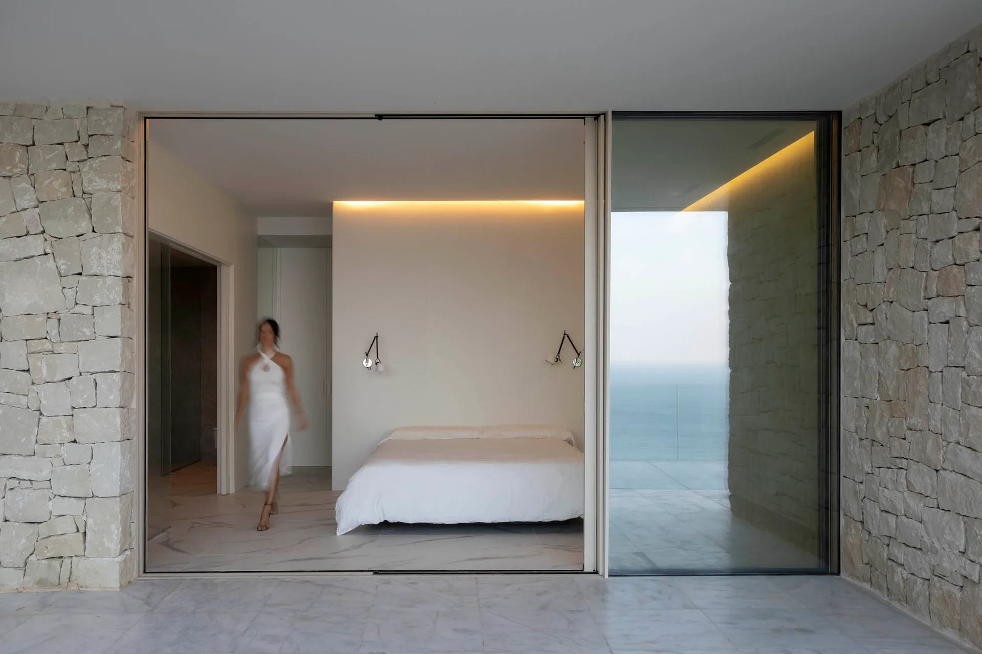 A minimalist bedroom with white walls covered in marble, featuring a single bed and a view of the ocean.