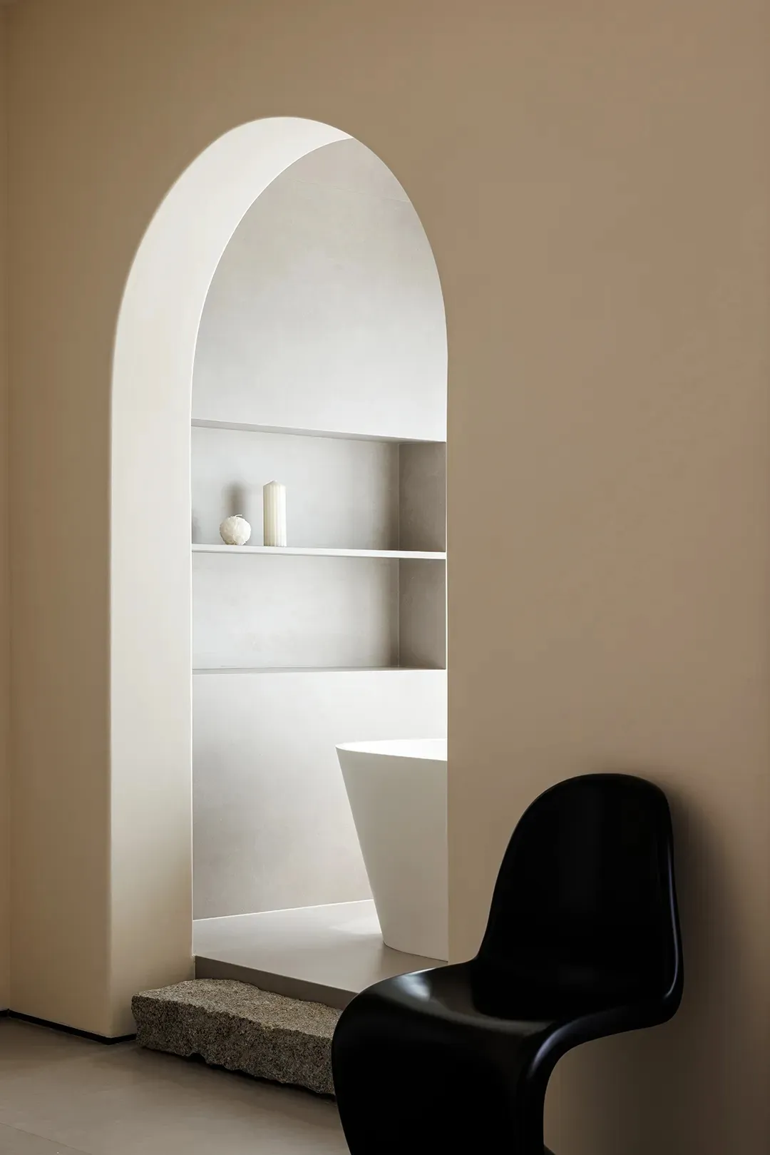 A minimalist bathroom with a white bathtub and a black chair against a beige wall, framed by an arched doorway and a stone st