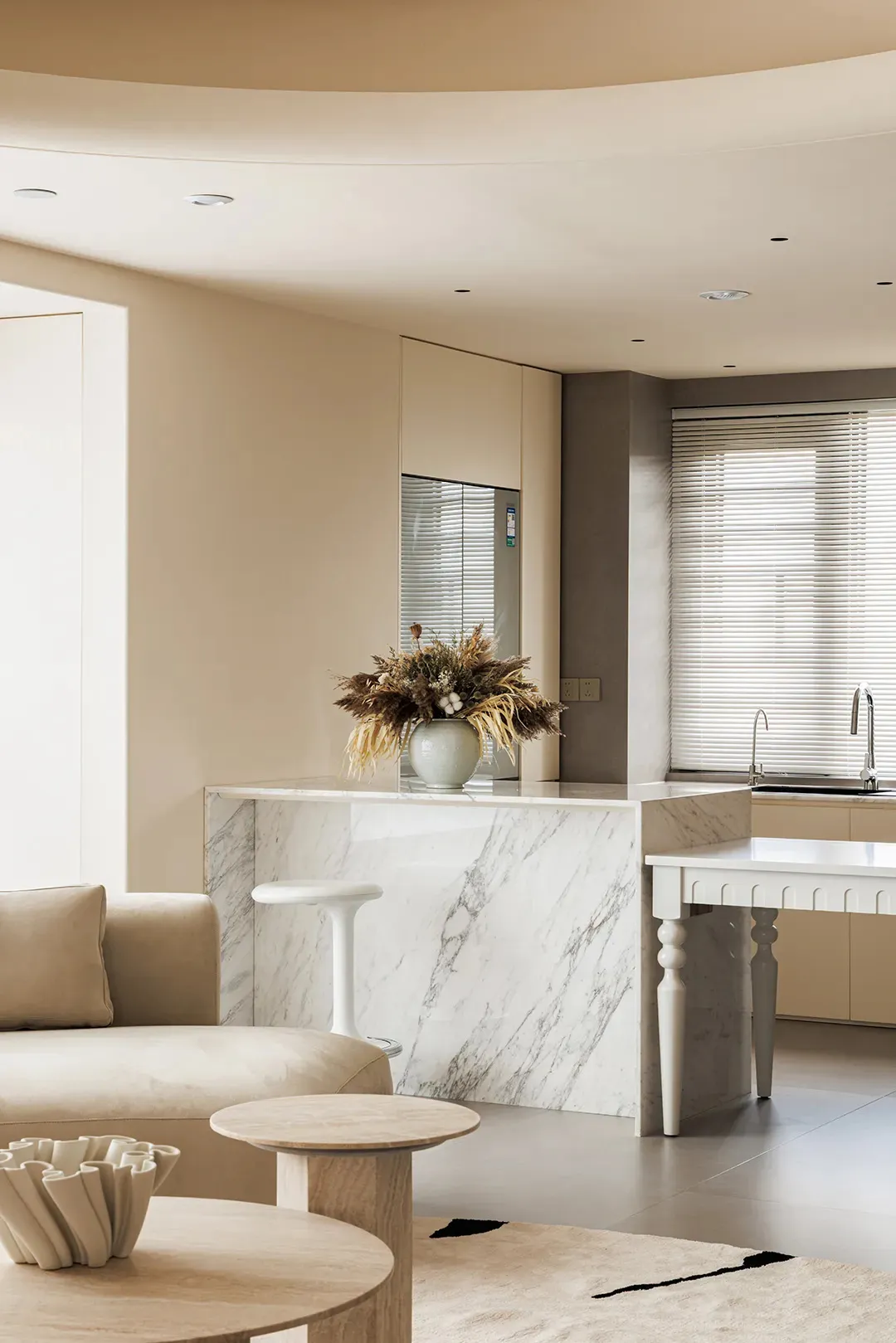 A modern minimalist kitchen featuring marble countertops and furniture, with a vase of dried flowers on the island.