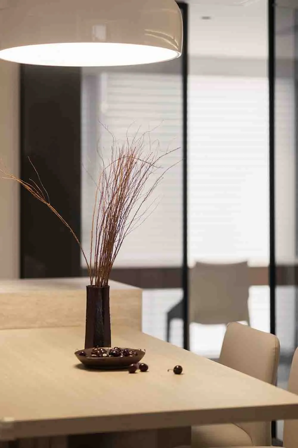 A minimalist dining room setup featuring a table with a decorative vase containing dried grasses and a bowl of dark stones.