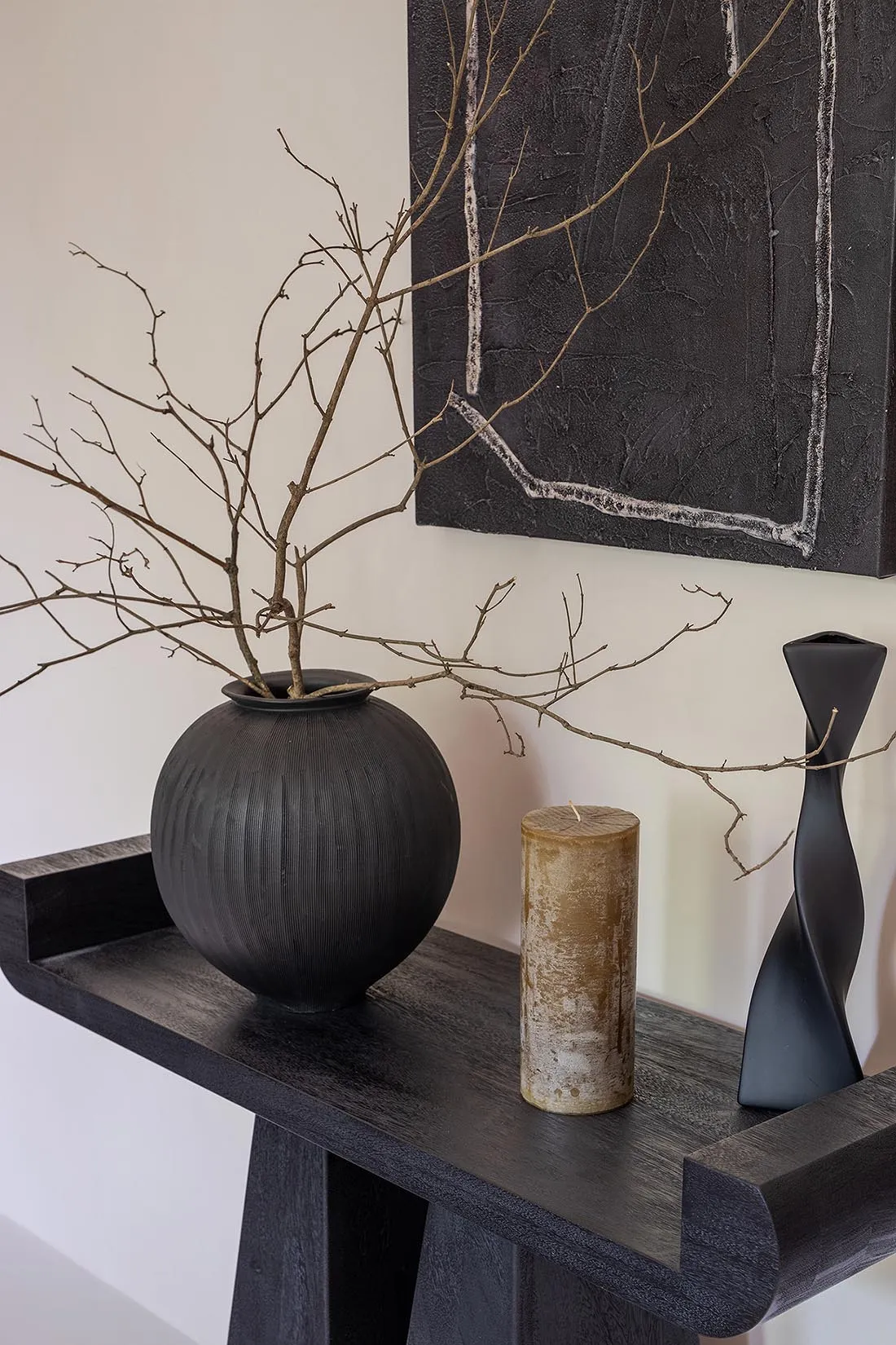 A black vase with dry branches in it, a cylindrical candle, and a black sculptural vase on a black stand in front of a black