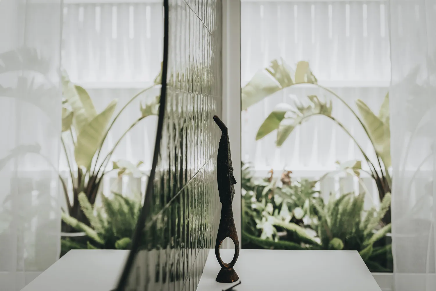 A minimalist metal sculpture on a white table in a well-lit room with green plants and white curtains.