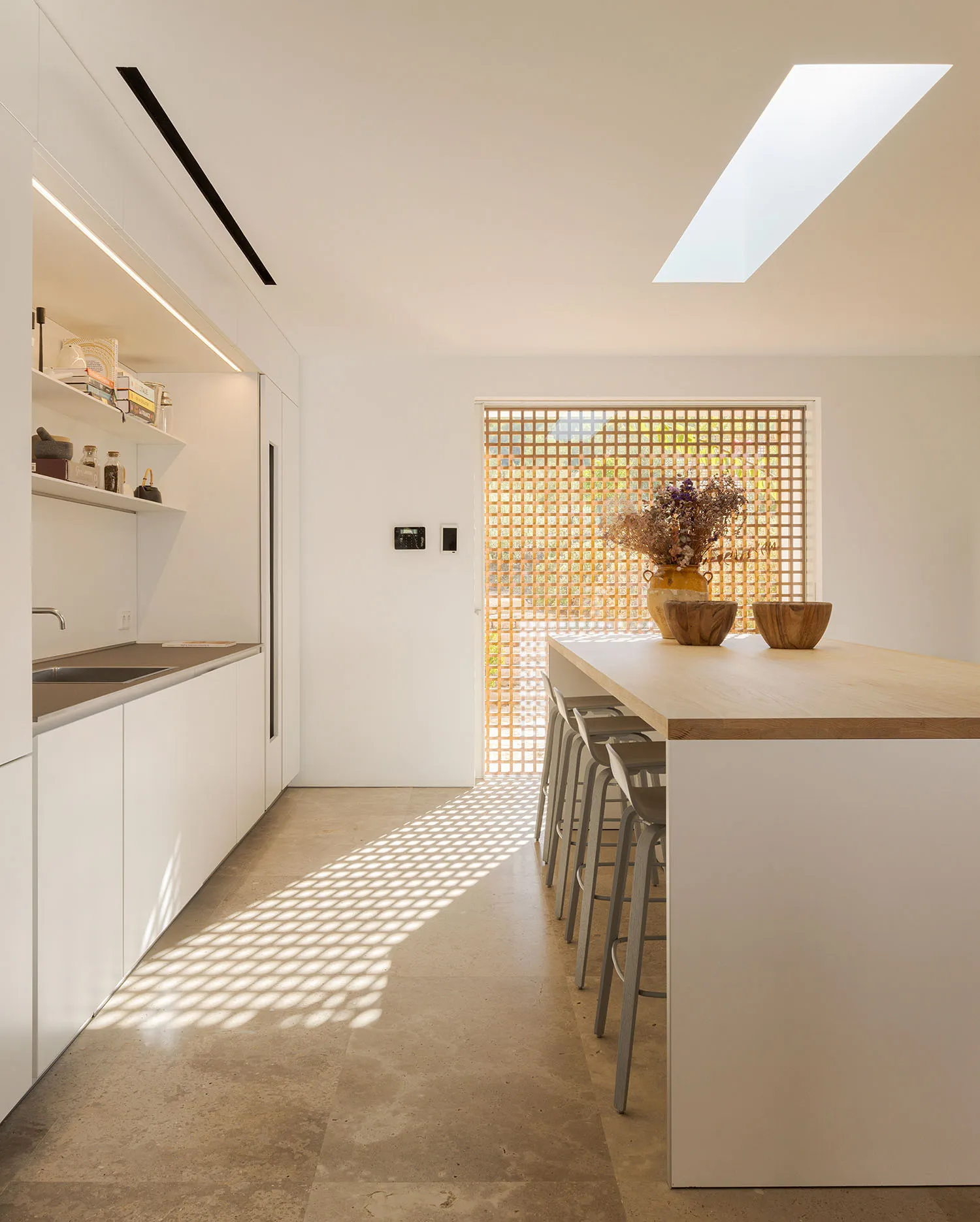 A modern minimalist kitchen with a wooden floor, featuring a long island with stools and a skylight.