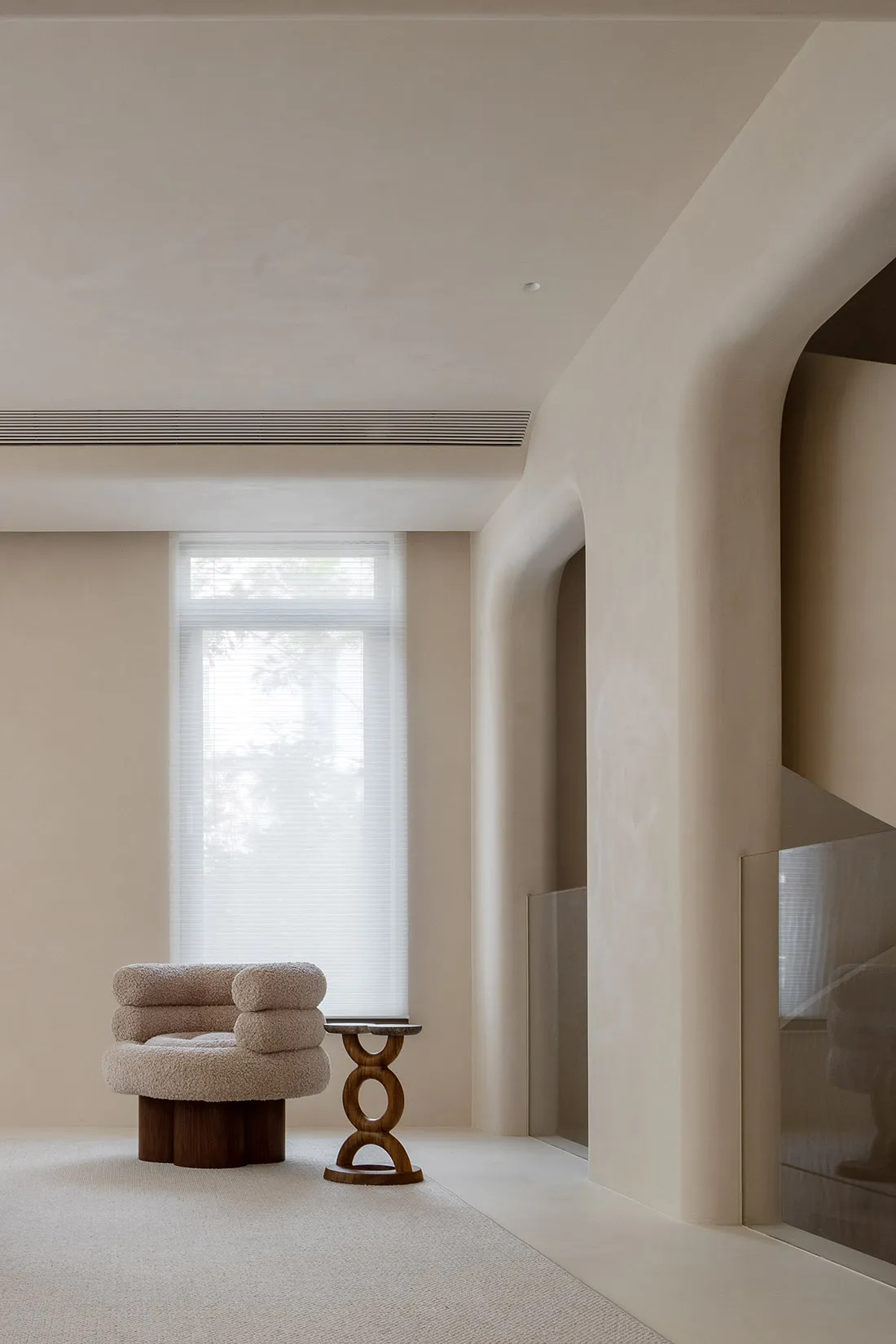 A minimalist living room with neutral-toned walls, a single plush armchair with pillows, a small wooden side table, and a lar