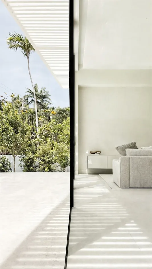 Modern minimalist living room with concrete flooring, white walls, and large windows overlooking palm trees.