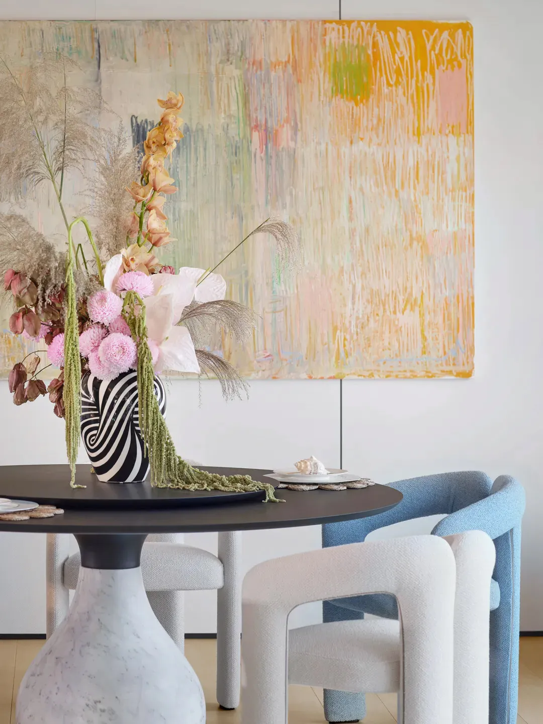 A modern dining room with a round table, blue upholstered chairs, and a floral arrangement in a vase with mixed media element