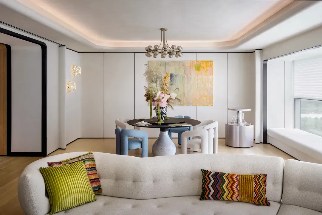 Modern living room with a white fabric sofa, colorful throw pillows, and a round dining table with a floral centerpiece.