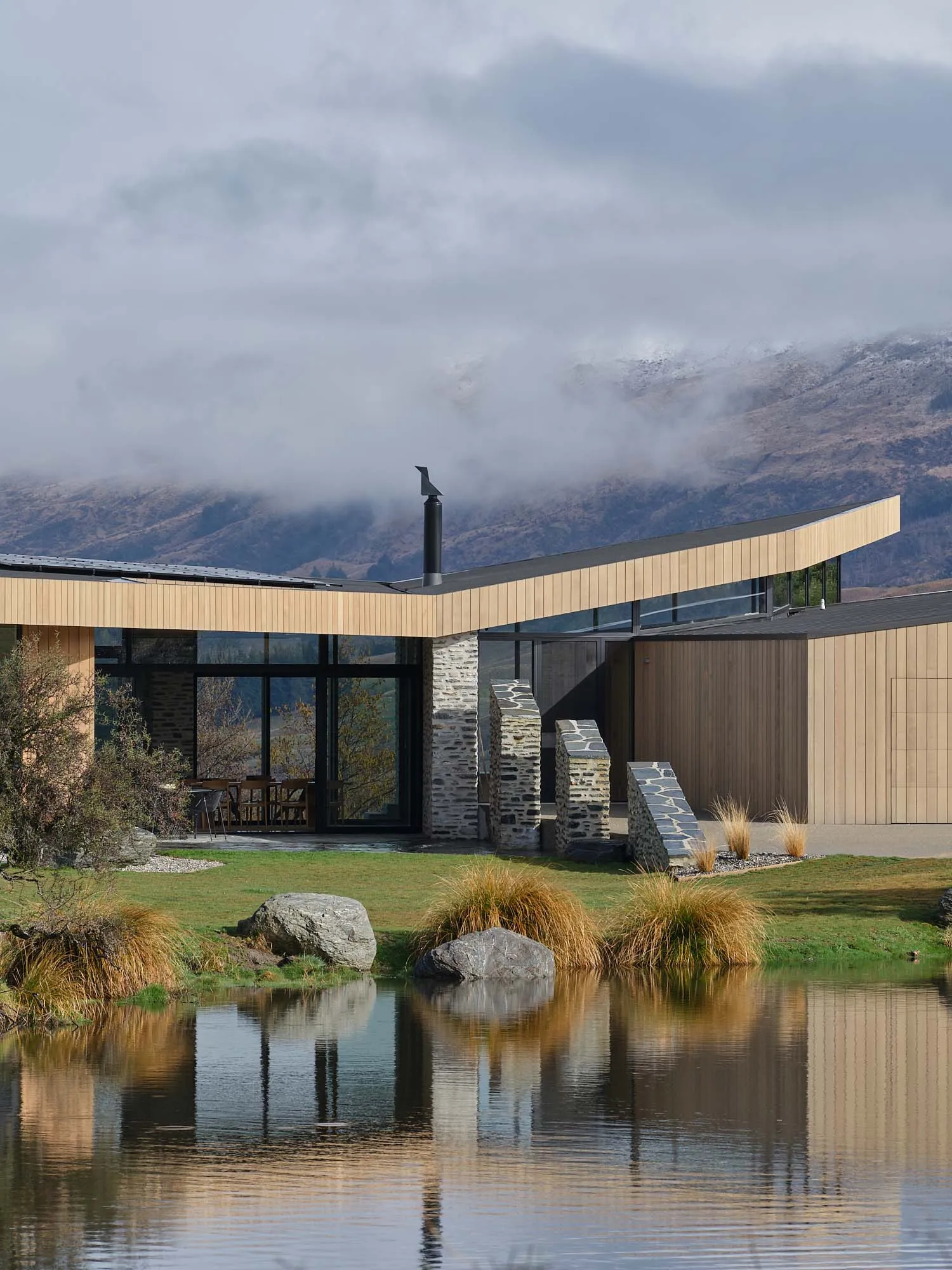 A modern house with a wooden exterior sits on the shore of a calm lake with mountains in the background.