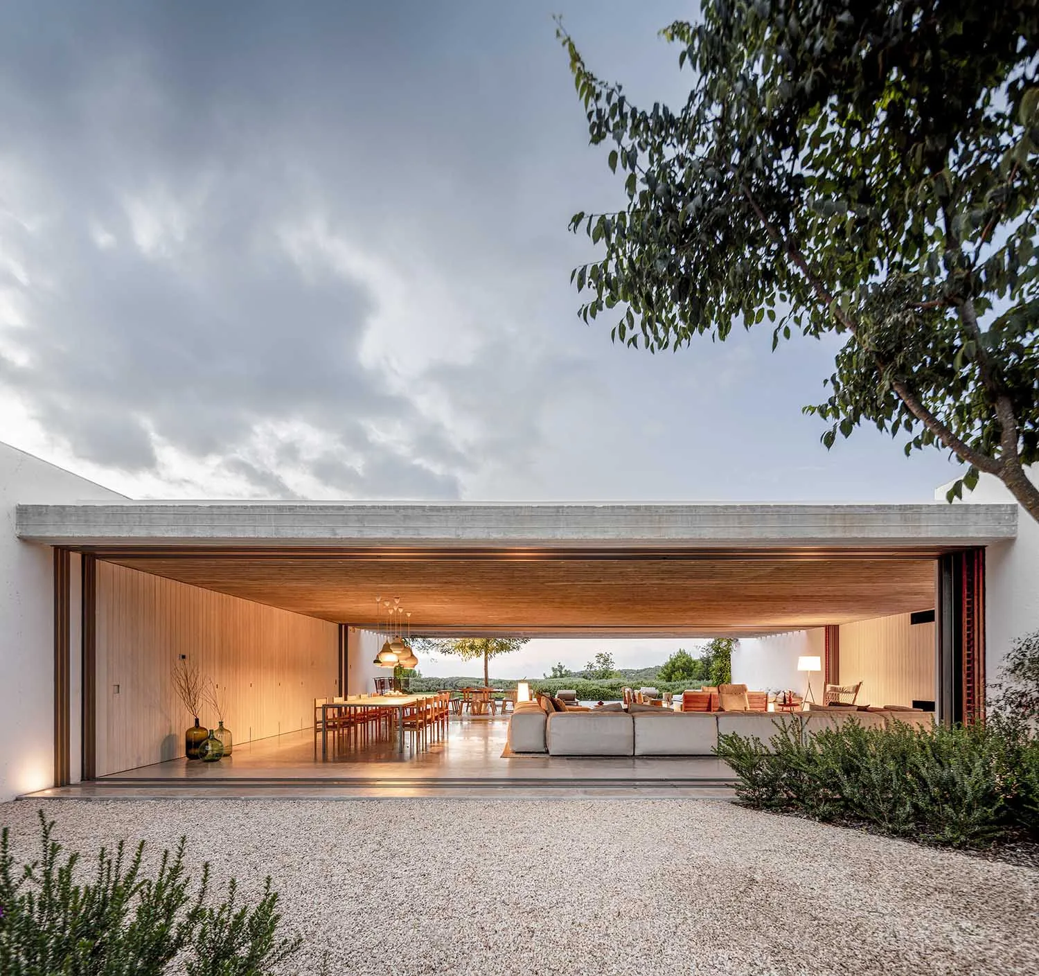A minimalist exterior living room with a wooden ceiling and floor, opening up to a gravel driveway and surrounding greenery.