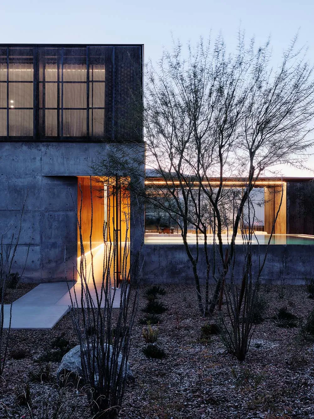 A modern house with a concrete exterior featuring large windows and a swimming pool at dusk.