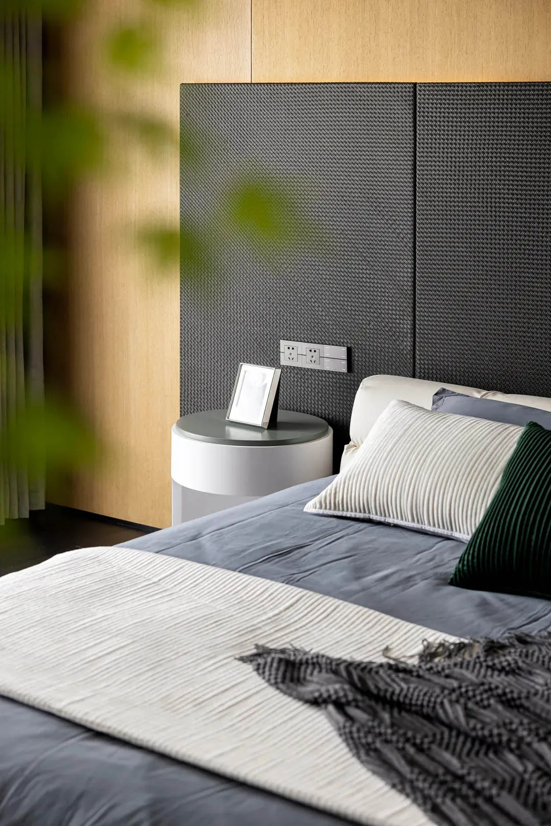 Modern minimalist bedroom featuring a textured headboard, grey blanket, green and white pillows, and grey bedframe with dark