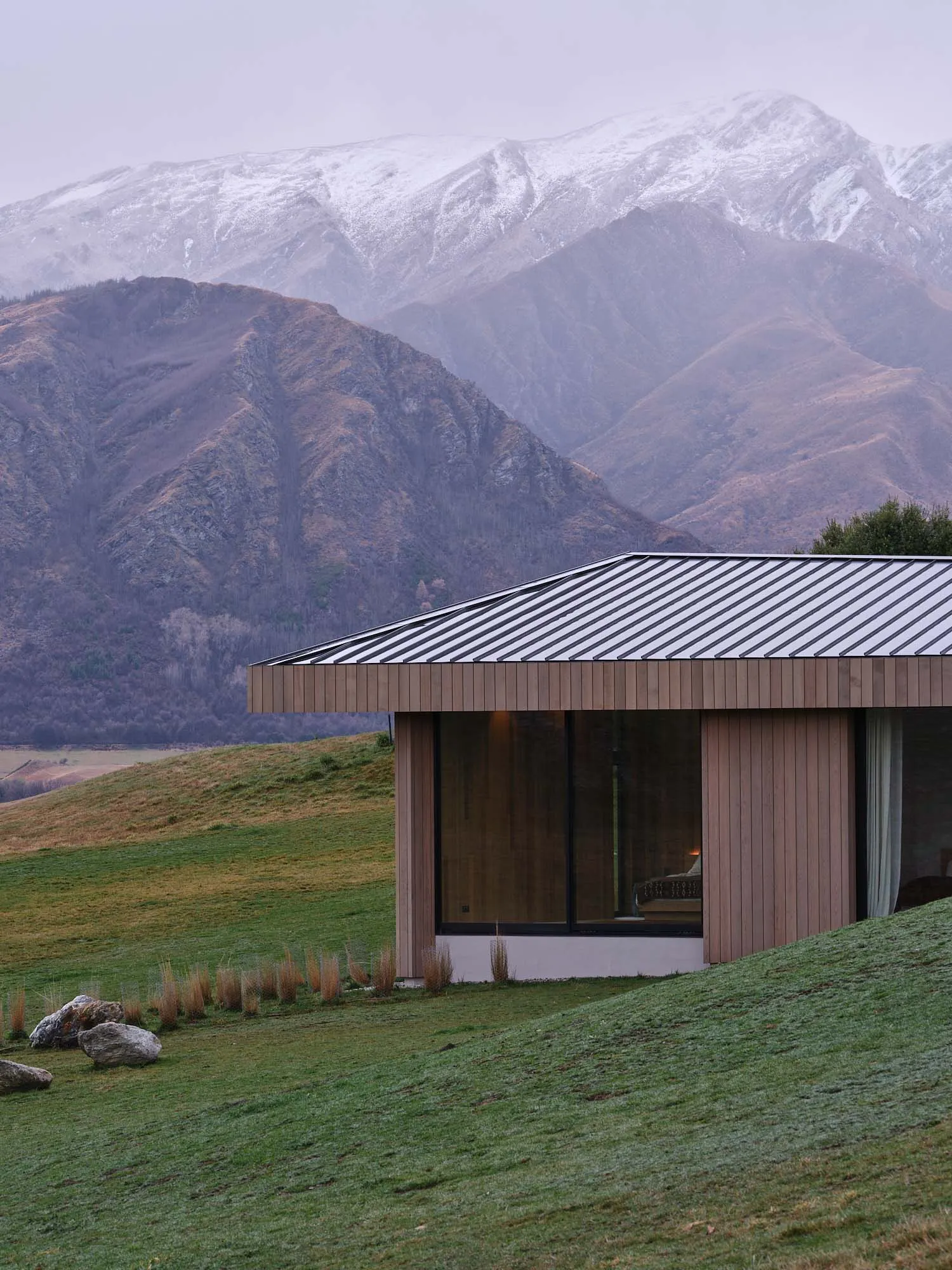 Modern minimalist residential exterior featuring a wooden façade with a metal roof, set against a backdrop of majestic snowy