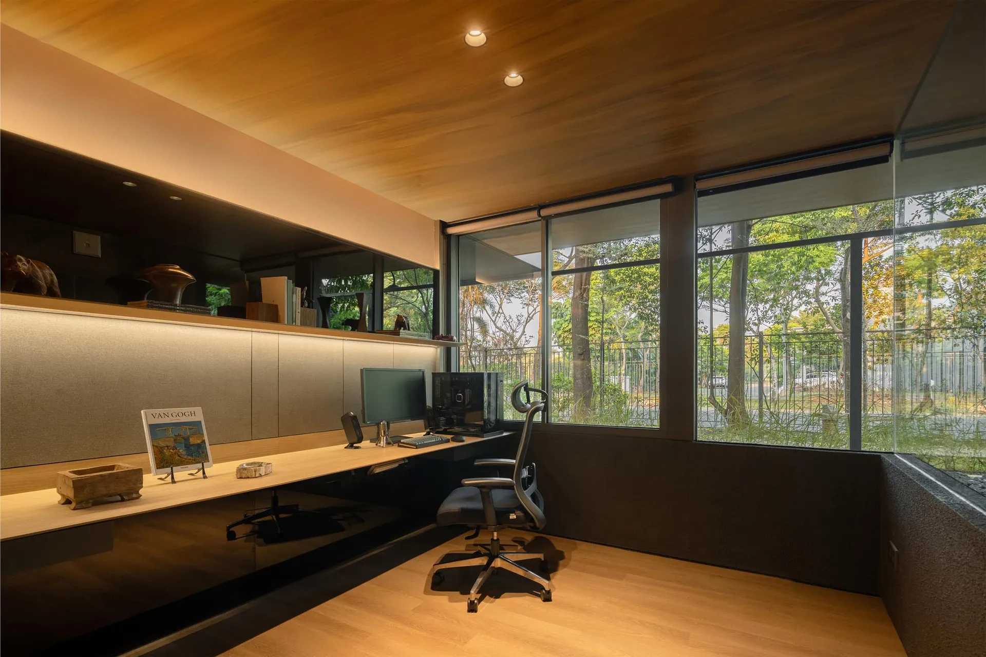 A minimalist office space featuring a desk, chair, and computer in a natural setting with large windows.