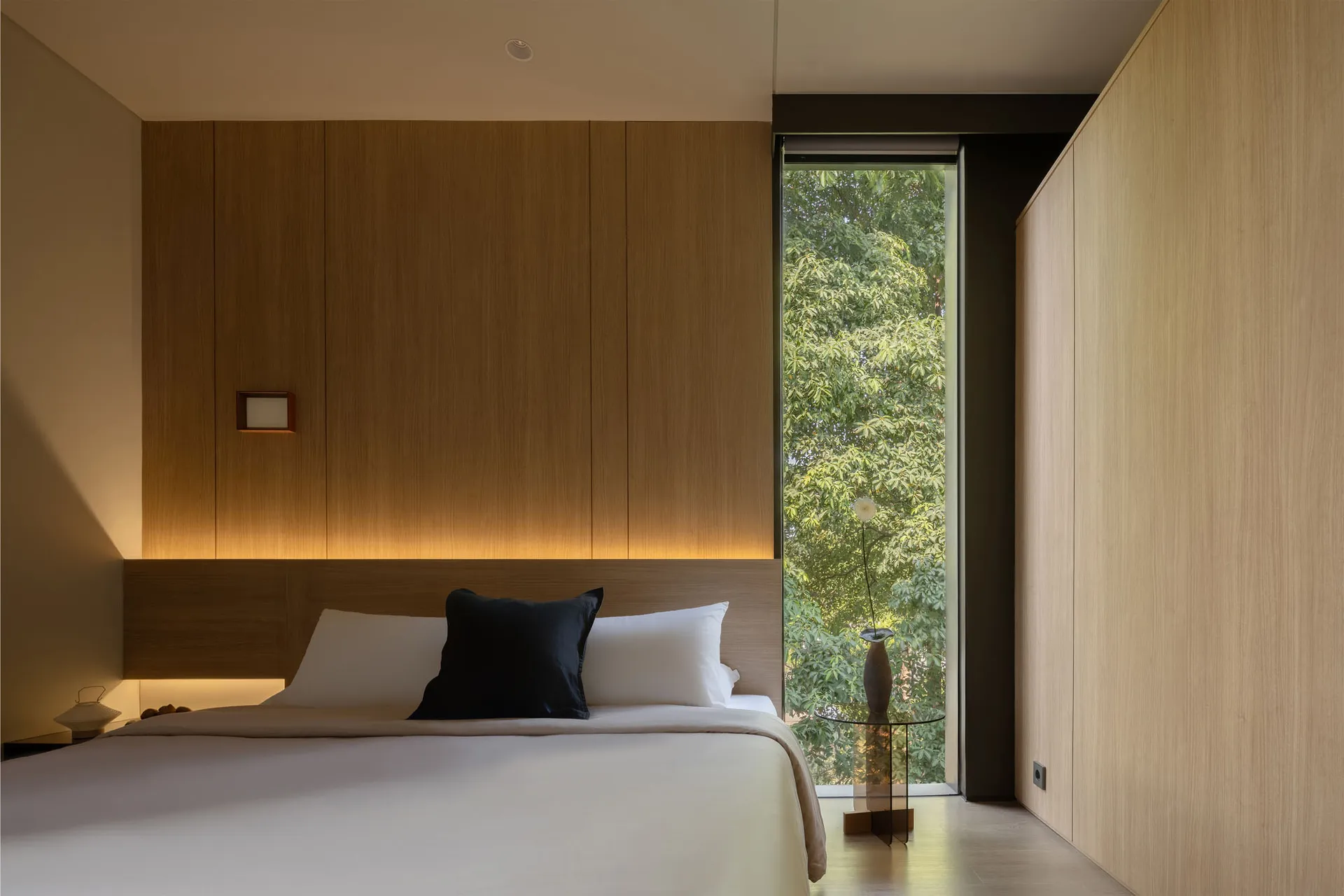 A modern bedroom featuring wooden paneling, a large window, and a bed with white sheets and black accent pillow.