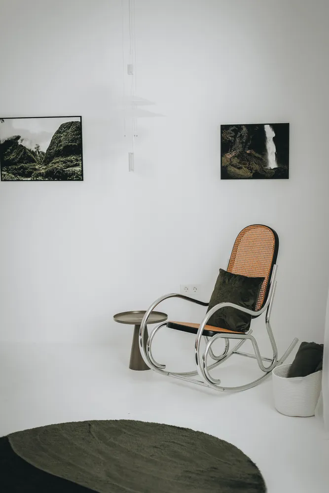 A minimalist living room featuring a metal rocking chair with a green cushion and a round rug on a white floor.
