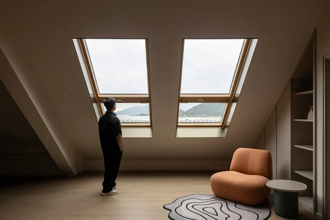 Man standing near skylights, looking out at mountains and sea, minimalist interior with orange chair and patterned rug.
