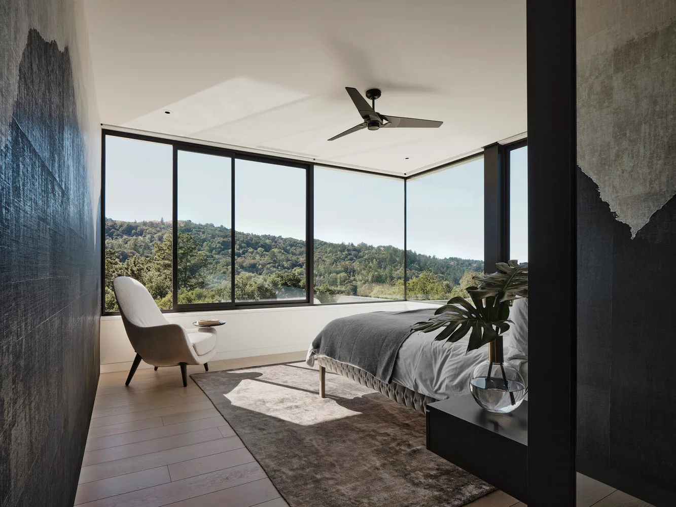A modern minimalist bedroom featuring a bed with grey sheets and a metal headboard, surrounded by large windows overlooking a