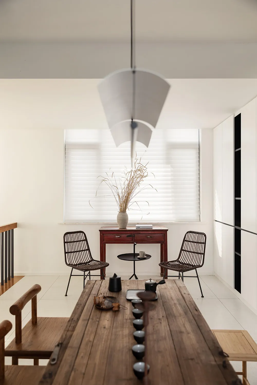 A minimalistic living room featuring a wooden table with black ceramic tea cups and a white vase with dried plants by the win