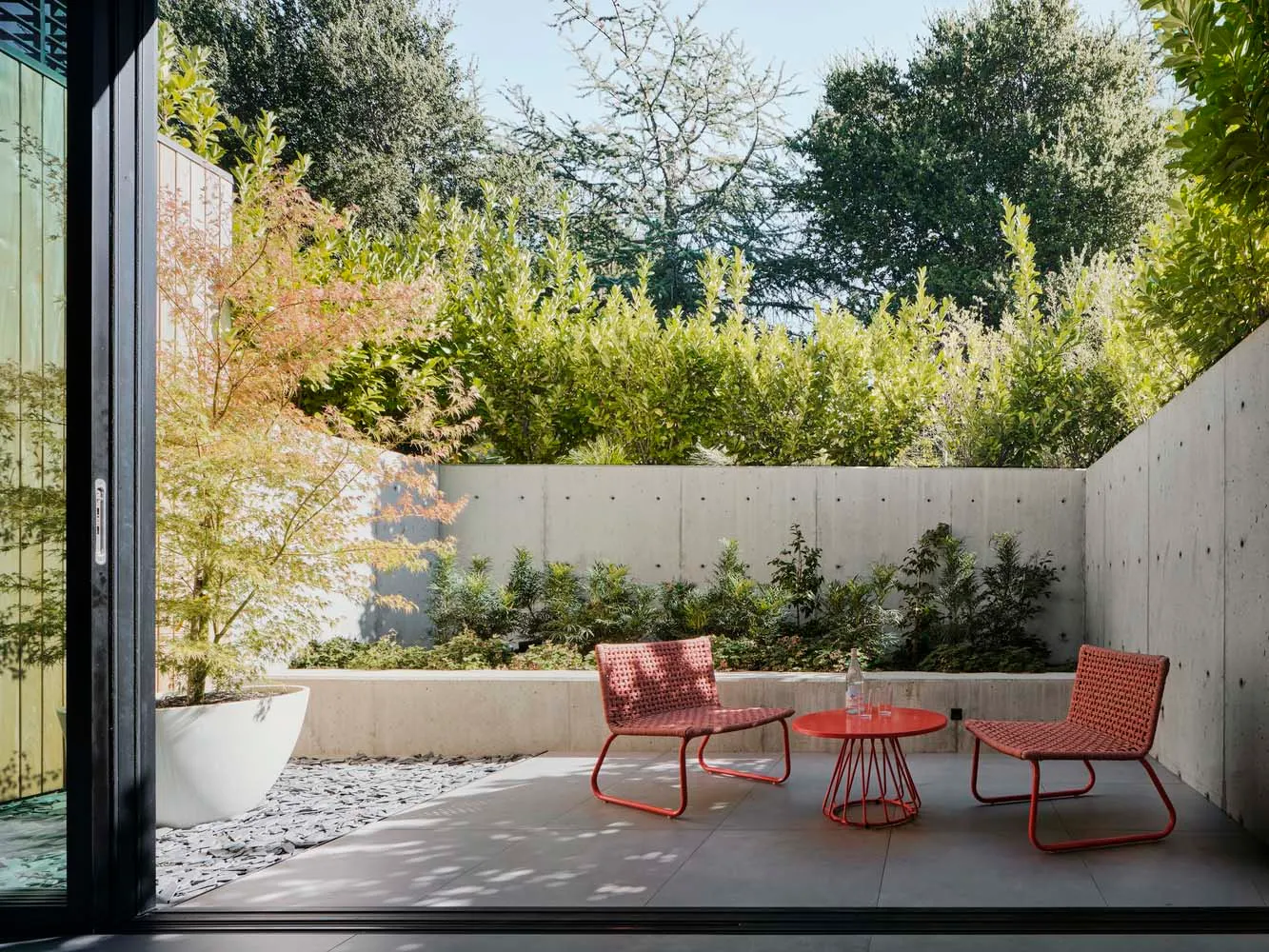 Minimalist outdoor living space with concrete walls, stone path, and red outdoor seating furniture.