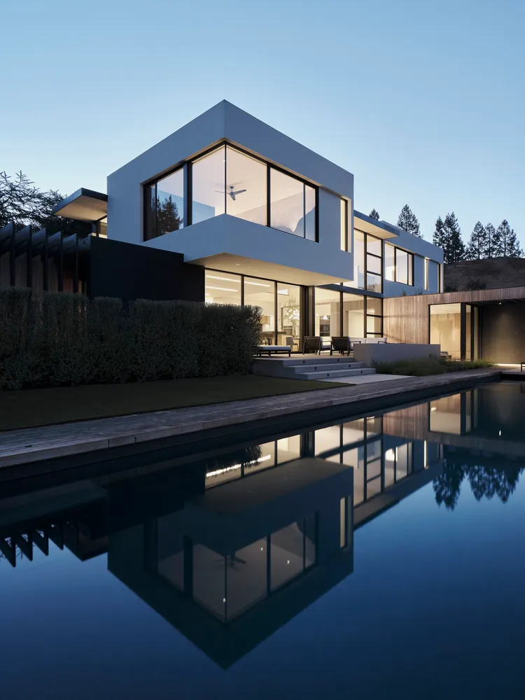 A modern house with a glass, concrete, and wooden exterior reflected in a still pool.